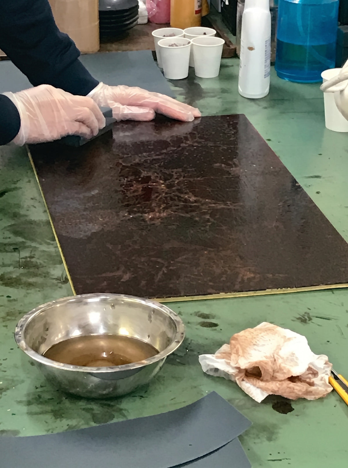 A craftsperson in gloves polishing a lacquered panel by hand, with a bowl of amber urushi lacquer beside it